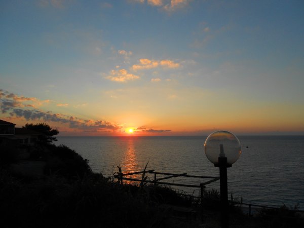 Tramonto sul mare con cielo nuvoloso, lampione a globo in primo piano.