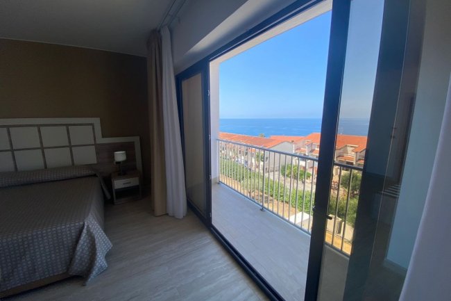 Camera con letto e grande finestra che si affaccia su un balcone con vista sul mare.