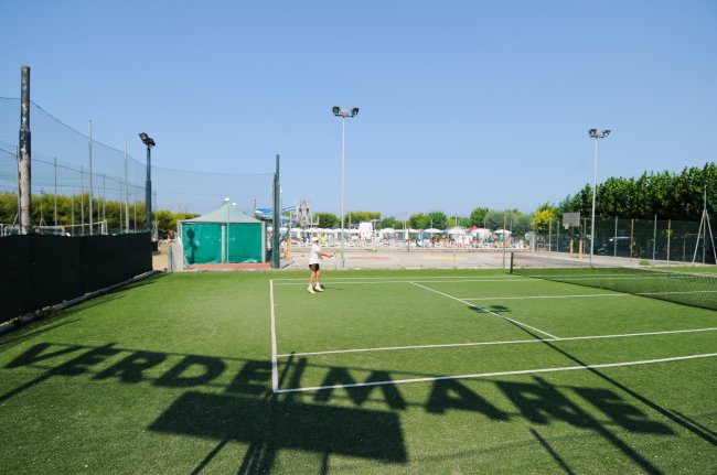 Campo da tennis in erba sintetica con una persona che gioca, sotto il cielo blu.