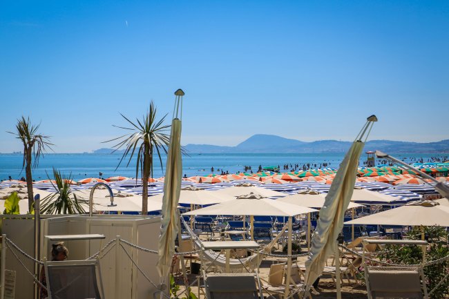 Spiaggia con ombrelloni bianchi, arancioni e blu. Palme e montagne all'orizzonte.