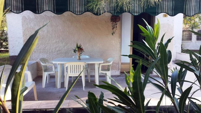 Veranda con tavolo bianco e sedie in plastica, coperta da un tendone verde, circondata da piante.