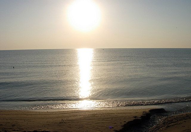 Mare calmo e sole al tramonto che si riflette sull'acqua, con una spiaggia in primo piano.