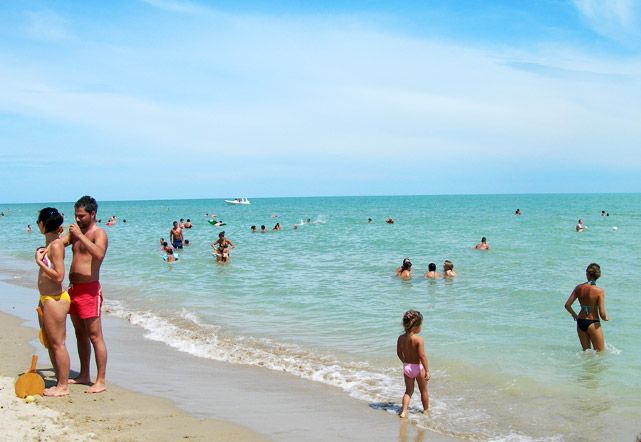 Spiaggia affollata con persone nel mare e sulla riva, sotto un cielo sereno.