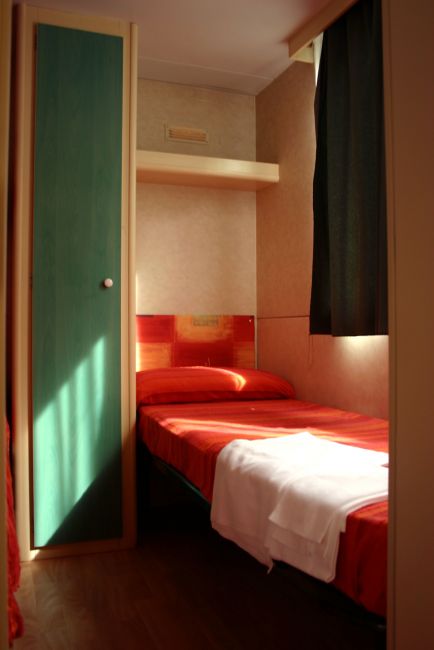 Una piccola stanza con letto singolo, lenzuola rosse e armadio verde chiaro.