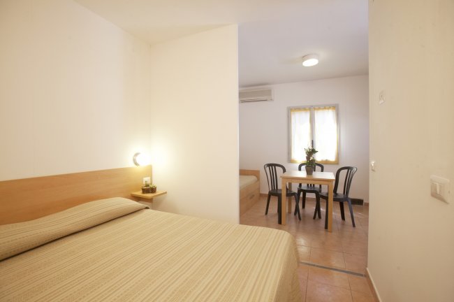 Interni di una stanza con letto matrimoniale, tavolo con sedie e finestra.