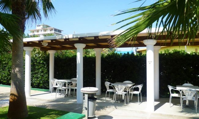 Zona pranzo all'aperto con tavoli e sedie sotto un pergolato.