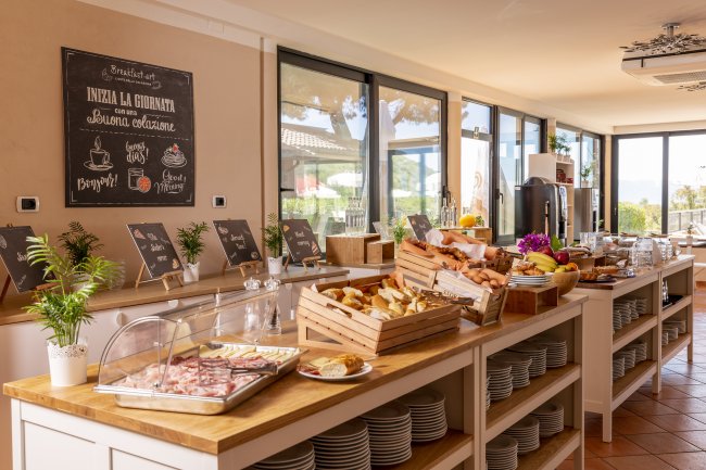 Buffet di colazione con pane, affettati, frutta e cartelli. Grandi finestre sullo sfondo.