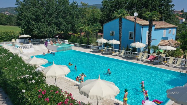Piscina all'aperto con sdraio e ombrelloni bianchi, circondata da vegetazione e un edificio blu.