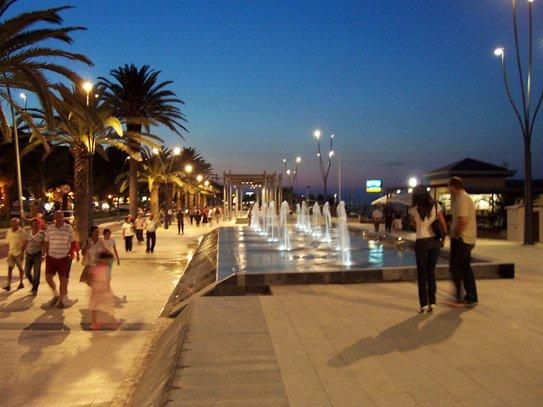 Passeggiata serale con fontane illuminate accanto a palme.