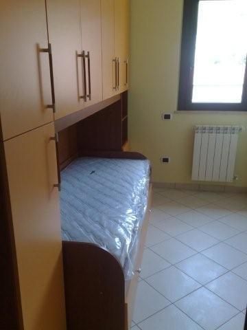 Letto singolo con materasso avvolto in plastica, inserito tra armadi e una finestra.