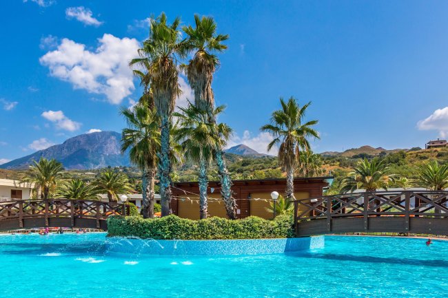 Piscina con fontane, palme e un ponte di legno, sfondo montuoso sotto un cielo azzurro.