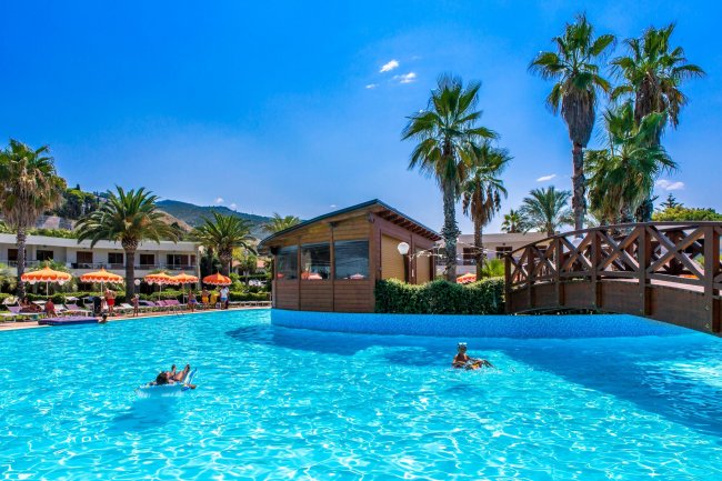 Piscina con palme, un ponte in legno e persone che nuotano sotto un cielo blu.