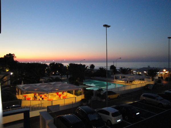 Vista serale su un patio con tavolini e vista mare, auto parcheggiate e cielo al tramonto.