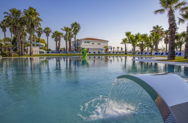 Piscina con fontana e palme, edificio bianco sullo sfondo.