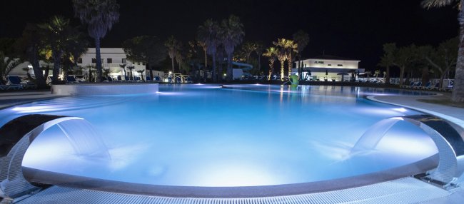 Piscina illuminata di sera con palme intorno e cascata d'acqua.