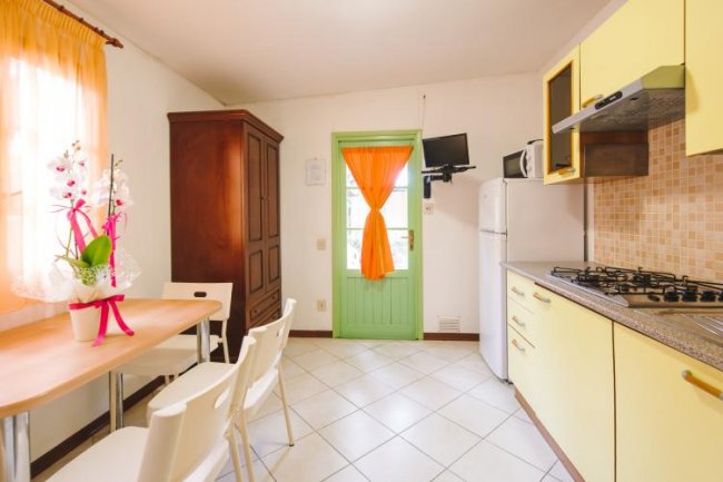 Cucina con porta verde, mobile alto in legno, tavolo con fiori e sedie bianche.