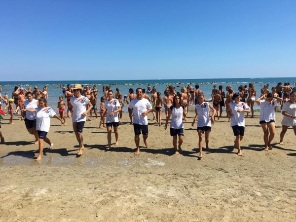Persone in fila sulla spiaggia partecipano ad attività, con il mare sullo sfondo.
