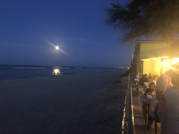 Una spiaggia di sera con luna piena e tavoli all’aperto illuminati.